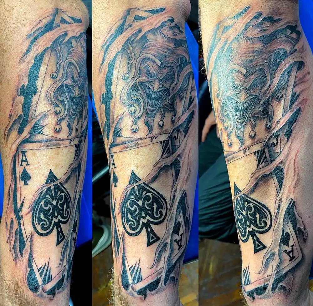 playing cards with an evil joker face and spade shredding thru skin black and grey by Power Dude of Dudes Tattoos, Bronx NY